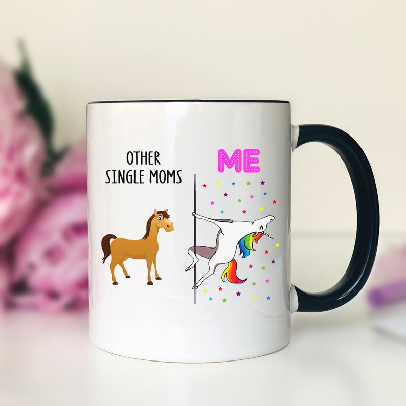 K&ouml;nnte beinhalten: Wei&szlig;e Keramik-Kaffeetasse mit schwarzem Henkel. Die Tasse hat eine schwarze Linie in der Mitte. Auf der linken Seite der Tasse steht der Text "Other Single Moms" mit einer Illustration eines braunen Pferdes. Auf der rechten Seite der Tasse steht der Text "ME" in Pink mit einer Illustration eines Einhorns, das eine Stange h&auml;lt.