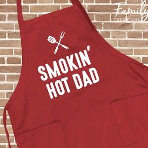 May include: A red apron featuring the text "SMOKIN' HOT DAD" in white, along with a crossed spatula and fork graphic. It has two front pockets and neck and waist ties. The apron is made of a durable fabric.