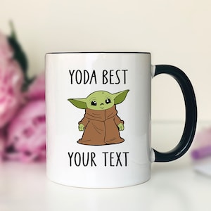 May include: White ceramic mug with a black handle and rim, featuring a green cartoon character with the text "YODA BEST YOUR TEXT". The character is wearing a brown cloak. The mug is likely for beverages.