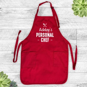 May include: A red apron with two front pockets and long ties. The apron features the text "Ashley's PERSONAL CHEF" in white, along with a crossed fork and spoon graphic. The apron is designed for cooking and food preparation.