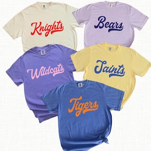 May include: Five short-sleeved t-shirts in cream, lavender, purple, yellow, and blue. Each shirt features a team name in a script font: Knights, Bears, Wildcats, Saints, and Tigers. The shirts are arranged in a casual display.
