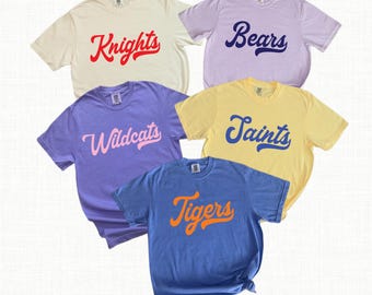 Puede incluir: Cinco camisetas de manga corta en crema, lavanda, morado, amarillo y azul. Cada camiseta presenta un nombre de equipo en fuente cursiva: Knights, Bears, Wildcats, Saints y Tigers. Las camisetas están dispuestas de forma informal.