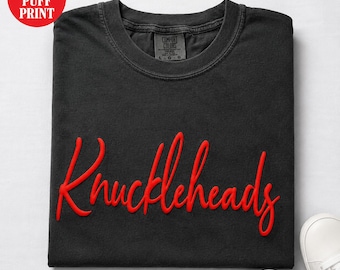 May include: A black crew neck t-shirt featuring the word Knuckleheads in a raised red puff print across the chest. The shirt has a Comfort Colors brand label on the inside collar. The design is a custom puff print style on a dark fabric base.