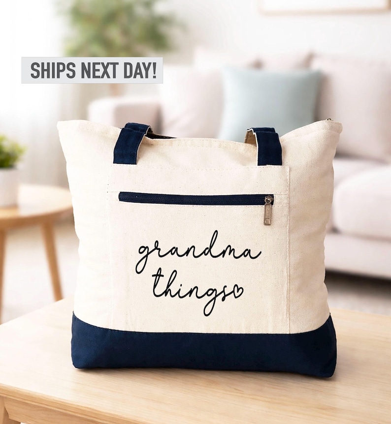 Pu&ograve; includere: Borsa tote color crema con dettagli blu navy. Presenta una tasca con cerniera e manici blu navy. Le parole "grandma things" sono scritte in corsivo sul davanti. La borsa &egrave; appoggiata su un tavolo di legno.
