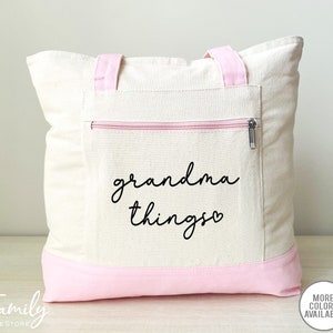 Pu&ograve; includere: Borsa tote color crema con dettagli rosa. La borsa presenta una tasca frontale con cerniera e le parole "grandma things" in corsivo nero. I manici e il fondo della borsa sono rosa.