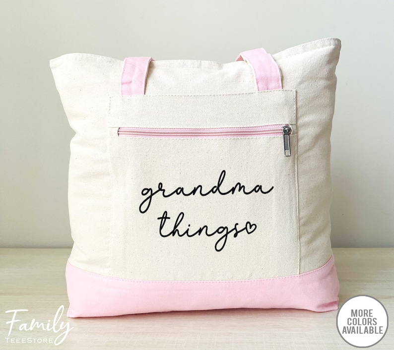Pu&ograve; includere: Borsa tote color crema con dettagli rosa. La borsa presenta una tasca frontale con cerniera e le parole "grandma things" in corsivo nero. I manici e il fondo della borsa sono rosa.
