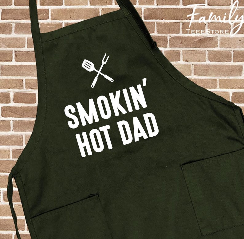 May include: Dark green apron with two front pockets and the text "SMOKIN' HOT DAD" in white. A white graphic of a spatula and fork is above the text. The apron has black straps and is against a brick wall background.