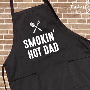 May include: Black apron with two front pockets and the text "SMOKIN' HOT DAD" in white. A white graphic of a spatula and fork crossed is above the text. The apron is designed for grilling and cooking.