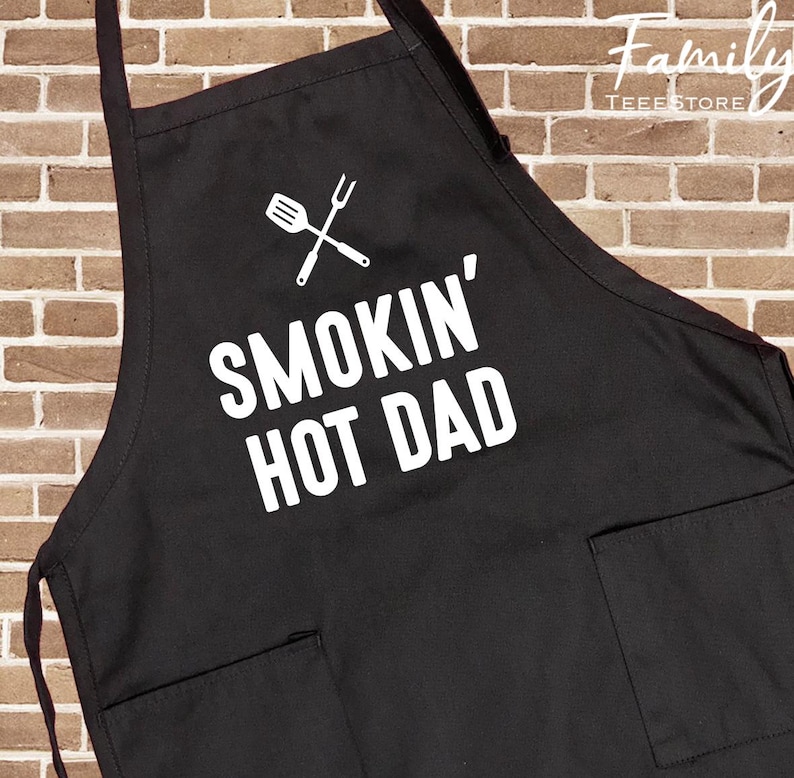 May include: Black apron with two front pockets and the text "SMOKIN' HOT DAD" in white. A white graphic of a spatula and fork crossed is above the text. The apron is designed for grilling and cooking.