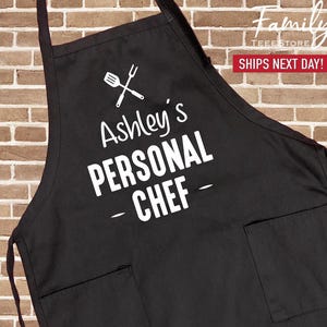 May include: Black apron with two front pockets. The apron features the text "Ashley's PERSONAL CHEF" in white, along with a graphic of a spatula and fork. The apron is designed for cooking and food preparation.