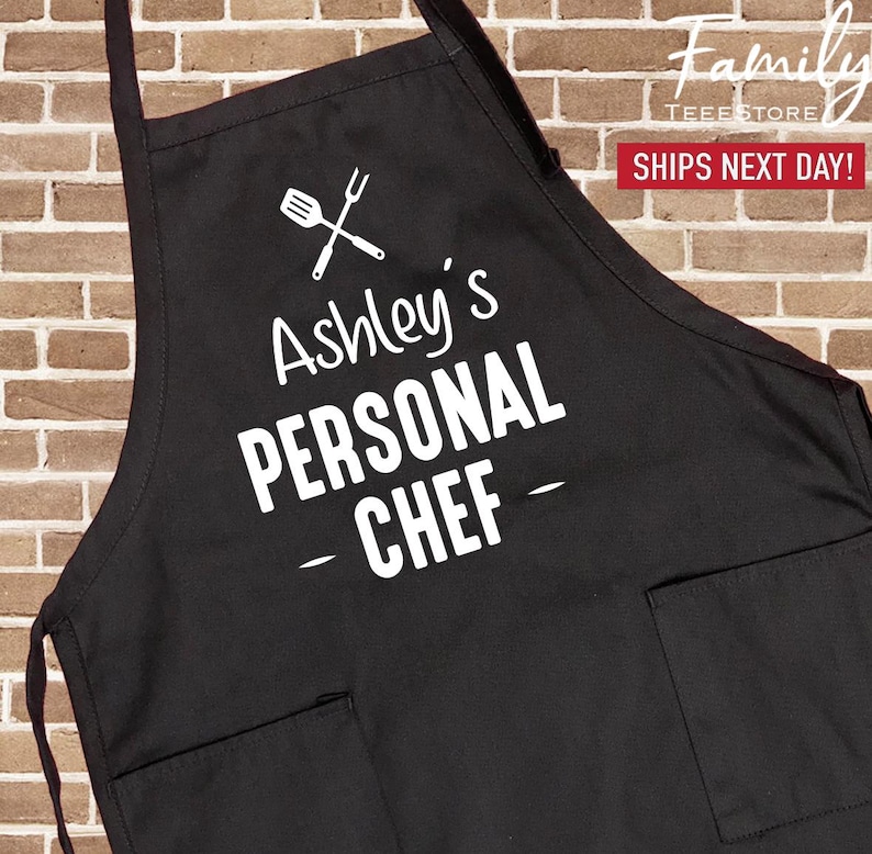 May include: Black apron with two front pockets. The apron features the text "Ashley's PERSONAL CHEF" in white, along with a graphic of a spatula and fork. The apron is designed for cooking and food preparation.