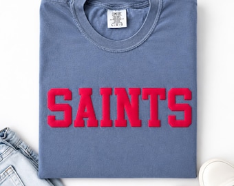 May include: A blue crew neck t-shirt with the word SAINTS printed in bold red block letters across the chest. The shirt features a Comfort Colors brand label at the inner neckline indicating a size large. The fabric has a soft, washed appearance.