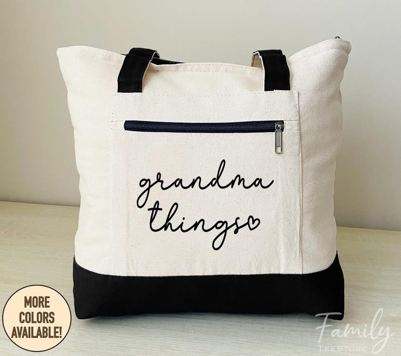 Pu&ograve; includere: Borsa tote color crema con dettagli neri, tra cui la base, i manici e una tasca con cerniera. La borsa presenta la scritta "grandma things" in corsivo. Un cerchio indica "MORE COLORS AVAILABLE!" in basso a sinistra.