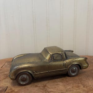 May include: A vintage brass car model with a classic design. The car has a detailed body, wheels, and a small windshield. The word "Corvette" is visible on the side. The car is set on a wooden surface.