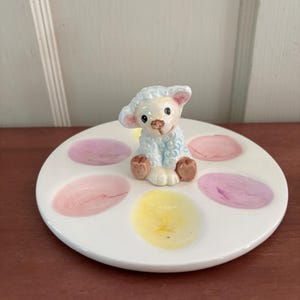May include: A white ceramic egg plate with six colorful egg-shaped paint splotches in pink, yellow, and purple. A small, light blue lamb figurine sits in the center. The lamb has brown hooves and a sweet expression.