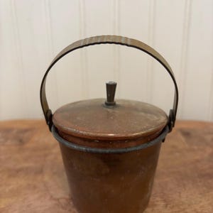 May include: A small, antique copper bucket with a lid and a handle. The bucket is a warm, reddish-brown color, with a matching lid and a curved handle. The handle is attached to the bucket with small metal brackets. The bucket is sitting on a wooden surface.