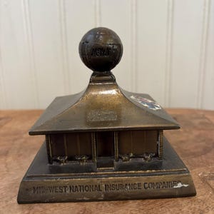 May include: A bronze-colored miniature building with a globe on top. The building has a pitched roof and the text "MID-WEST NATIONAL INSURANCE COMPANIES" is inscribed on the base. The globe has the word "NEW" on it.