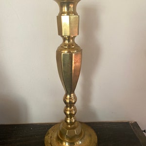 May include: A tall, brass candlestick with a wide base and a flared top. The candlestick has a geometric design with a series of octagonal and rounded sections. The brass has a warm, golden hue and shows signs of age.
