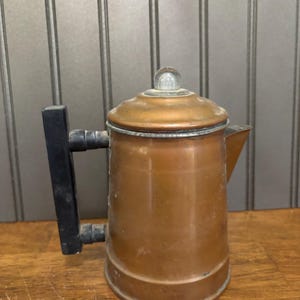 May include: Vintage copper coffee pot with a clear glass knob on the lid and a black handle. The pot has a spout and a rounded body. The copper has a warm, aged patina, suggesting it's been well-used.