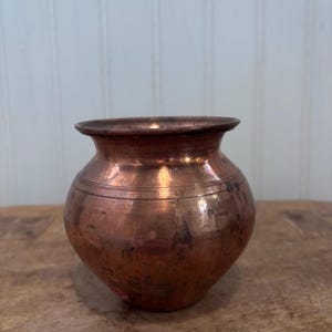 May include: A small, antique copper pot with a rounded body and a slightly flared rim. The pot has a rich, reddish-brown color with visible patina and subtle horizontal bands. It sits on a wooden surface against a white-paneled wall.