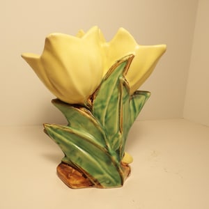 May include: A ceramic vase shaped like a large yellow tulip flower with green leaves at the base. The leaves feature dark brown accents along the edges and veins, providing a textured appearance to the stem and foliage supporting the yellow petals.