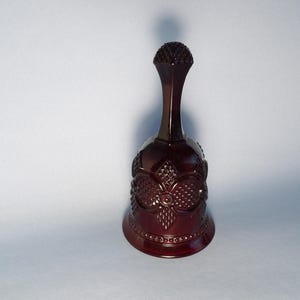 May include: A burgundy-colored bell with a detailed floral pattern. The bell has a long, slender handle and a textured top. The body of the bell features an embossed design with a series of diamond shapes and floral elements.