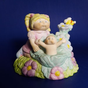 May include: A porcelain figurine of a Cabbage Patch Kid doll holding a baby Cabbage Patch Kid doll. The dolls are sitting in a bed of green leaves with pink flowers. A yellow and black bumblebee is flying above the flowers.