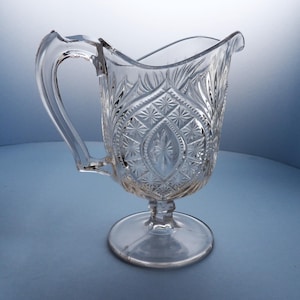 May include: A clear glass pitcher with a pedestal base and a cut glass design. The pitcher has a handle and a wide mouth.
