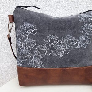 K&ouml;nnte beinhalten: Eine graue Canvas-Tragetasche mit braunem Lederboden. Die Tasche hat ein wei&szlig;es Blumenmuster.