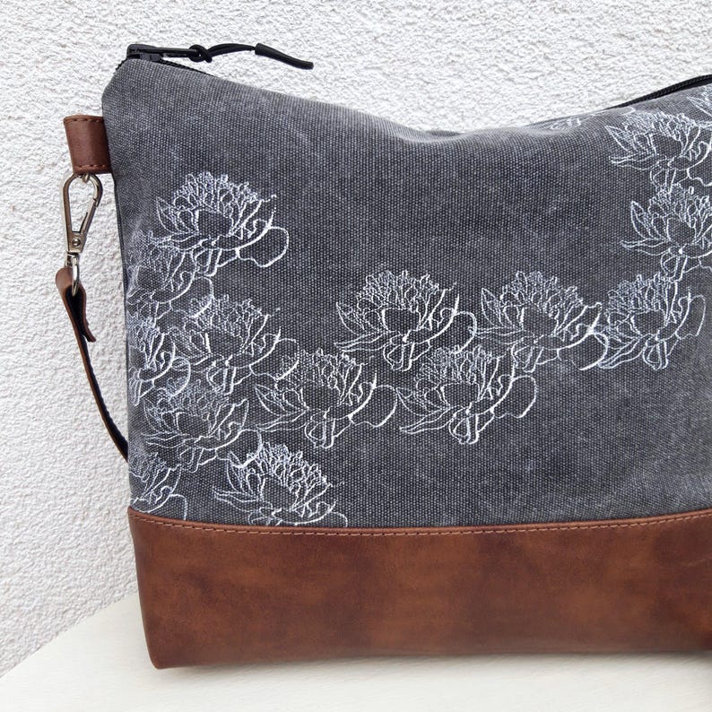 K&ouml;nnte beinhalten: Eine graue Canvas-Tragetasche mit braunem Lederboden. Die Tasche hat ein wei&szlig;es Blumenmuster.