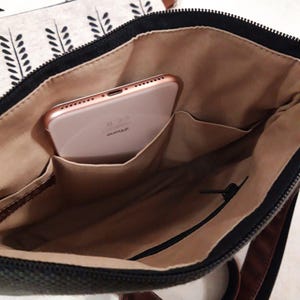 K&ouml;nnte beinhalten: Eine schwarze Canvas-Tasche mit einem braunen Lederriemen. Die Tasche ist offen und ein ros&eacute;goldenes iPhone befindet sich darin. Die Tasche hat eine Rei&szlig;verschlusstasche und ein braunes Futter.