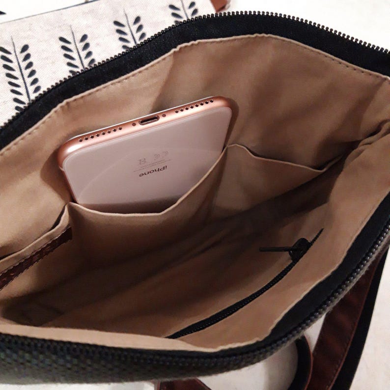 K&ouml;nnte beinhalten: Eine schwarze Canvas-Tasche mit einem braunen Lederriemen. Die Tasche ist offen und ein ros&eacute;goldenes iPhone befindet sich darin. Die Tasche hat eine Rei&szlig;verschlusstasche und ein braunes Futter.