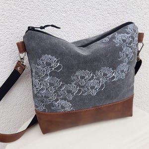 K&ouml;nnte beinhalten: Eine graue Canvas-Tragetasche mit braunem Lederboden. Die Tasche hat ein wei&szlig;es Blumenmuster und einen Rei&szlig;verschluss.