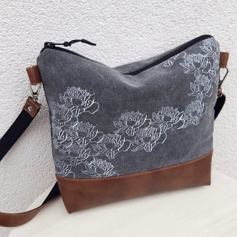 K&ouml;nnte beinhalten: Eine graue Canvas-Tragetasche mit braunem Lederboden. Die Tasche hat ein wei&szlig;es Blumenmuster und einen Rei&szlig;verschluss.