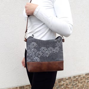 K&ouml;nnte beinhalten: Eine graue Canvas-Schultertasche mit braunem Lederboden und wei&szlig;er Blumenstickerei. Die Tasche hat einen langen verstellbaren Riemen.