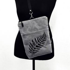 May include: Gray crossbody bag with a black fern leaf design. The bag has a front pocket and a black adjustable strap. The bag is displayed on a black mannequin.