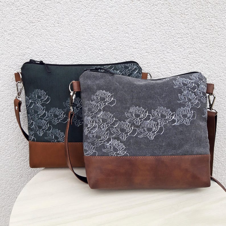 K&ouml;nnte beinhalten: Zwei graue Canvas-Umh&auml;ngetaschen mit wei&szlig;en Blumenmustern. Die Taschen haben braune Lederakzente und verstellbare Tr&auml;ger.