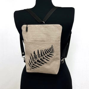 May include: A beige crossbody bag with a black fern leaf design. The bag has a front pocket, a zippered top, and adjustable black straps. The bag is displayed on a black mannequin.