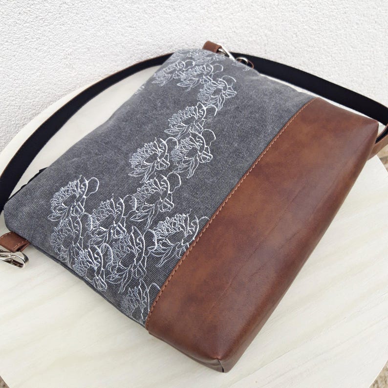 K&ouml;nnte beinhalten: Eine graue Canvas-Umh&auml;ngetasche mit braunem Lederboden. Die Tasche hat ein wei&szlig;es Blumenmuster, das auf der Vorderseite gestickt ist.
