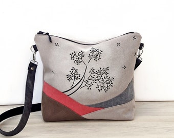 May include: A beige canvas crossbody bag with a black strap and a black tree design. The bag has a red, brown, and gray patchwork design.