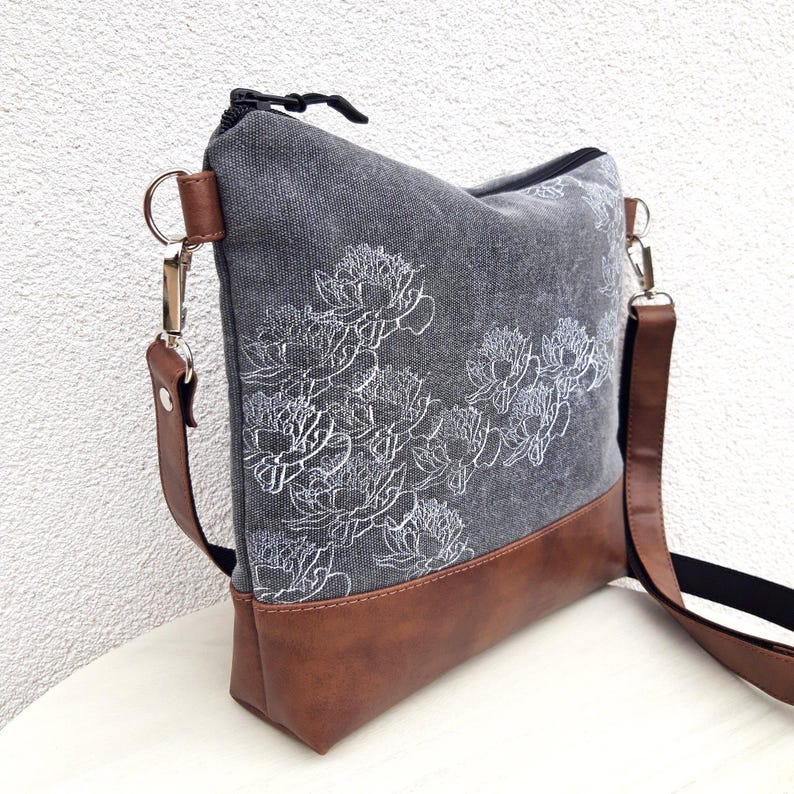 K&ouml;nnte beinhalten: Eine graue Canvas-Tragetasche mit braunem Lederboden und einem wei&szlig;en Blumendesign. Die Tasche hat einen langen verstellbaren Riemen.