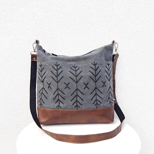 May include: Gray canvas shoulder bag with a brown leather base and strap. The bag features a black geometric tree pattern and a black adjustable strap. The bag has a zippered top and a front pocket.