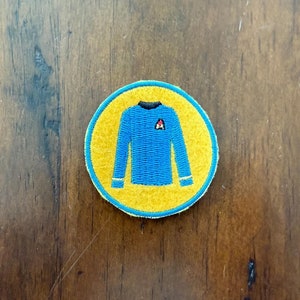 Puede incluir: Un parche redondo bordado de color amarillo con una camisa azul de manga larga con un símbolo delta rojo en la parte delantera.