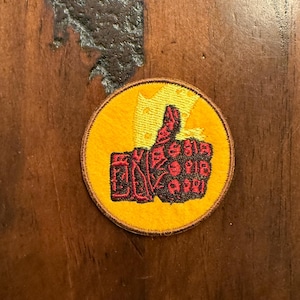 Può includere: Una toppa rotonda gialla con un disegno a pollice in su. Il pollice in su è rosso con dettagli neri, su sfondo giallo. La toppa ha un bordo giallo ed è probabilmente da stirare o cucire.