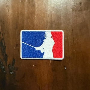 Puede incluir: Un parche bordado blanco con fondo azul y rojo. El parche presenta la silueta de un esgrimista sosteniendo un florete.