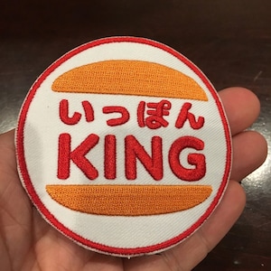 Może przedstawiać: Okrągła, biała naszywka haftowana z czerwoną obwódką. Naszywka przedstawia żółtą bułkę do hamburgera z napisem "いっぽん KING" w kolorze czerwonym.
