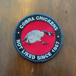 May include: A round, black embroidered patch with a red centre. The patch features a white goose with black eyes and a black beak. The text "COBRA CHICKENS NOT LIKED SINCE 1867" is embroidered in white around the edge of the patch.