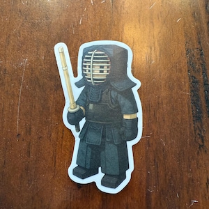Puede incluir: Una pegatina con una ilustración estilizada de un practicante de Kendo con armadura negra, sosteniendo una espada de madera. La figura lleva un casco con una rejilla protectora. La pegatina tiene un borde blanco y está sobre un fondo de madera.