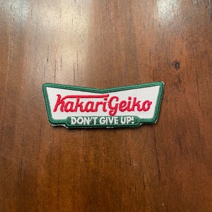 Peut inclure: Un écusson brodé blanc et vert avec le texte "Hakari Geiko" en rouge et "Don't Give Up!" en noir.