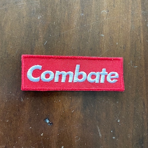 May include: Red rectangular patch with white embroidered text that reads "Combate".